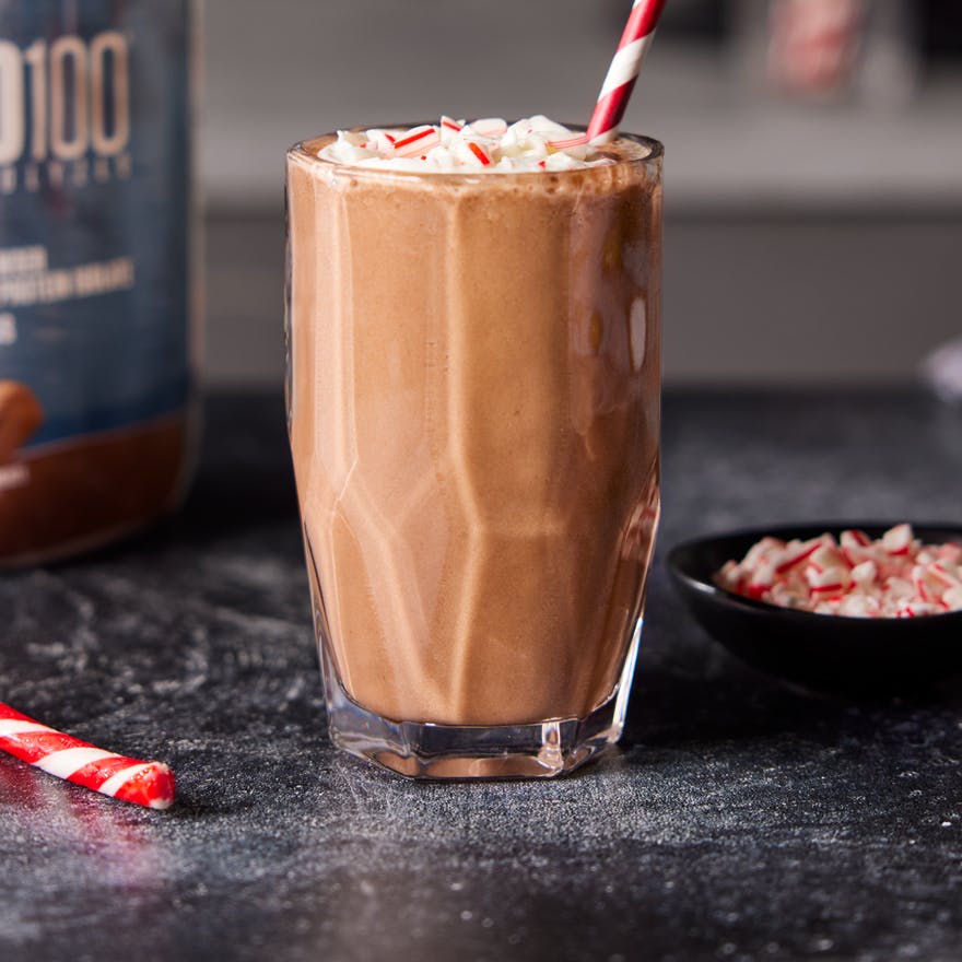 Chocolate Peppermint Protein Shake in a glass with festive straw. A tub of Dymatize and candy canes are nearby.