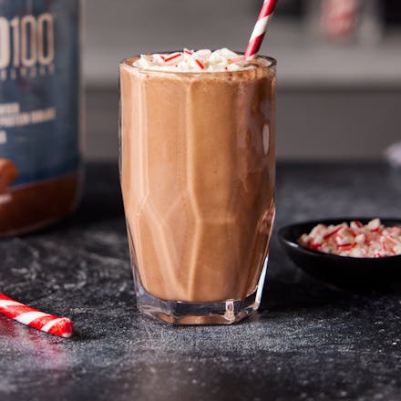 Chocolate Peppermint Protein Shake in a glass with festive straw. A tub of Dymatize and candy canes are nearby.