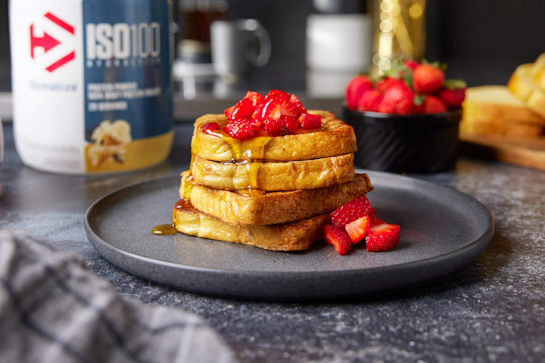 A stack of french toast covered in syrup and strawberries. A tub of Dymatize powder is nearby.
