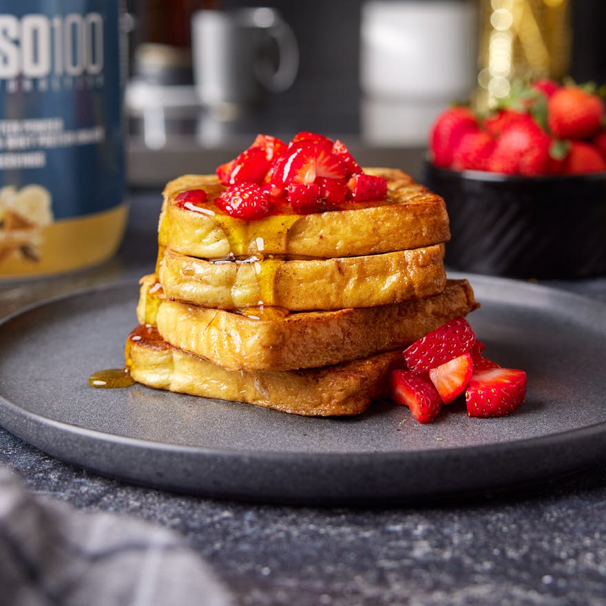 A stack of french toast covered in syrup and strawberries. A tub of Dymatize powder is nearby.