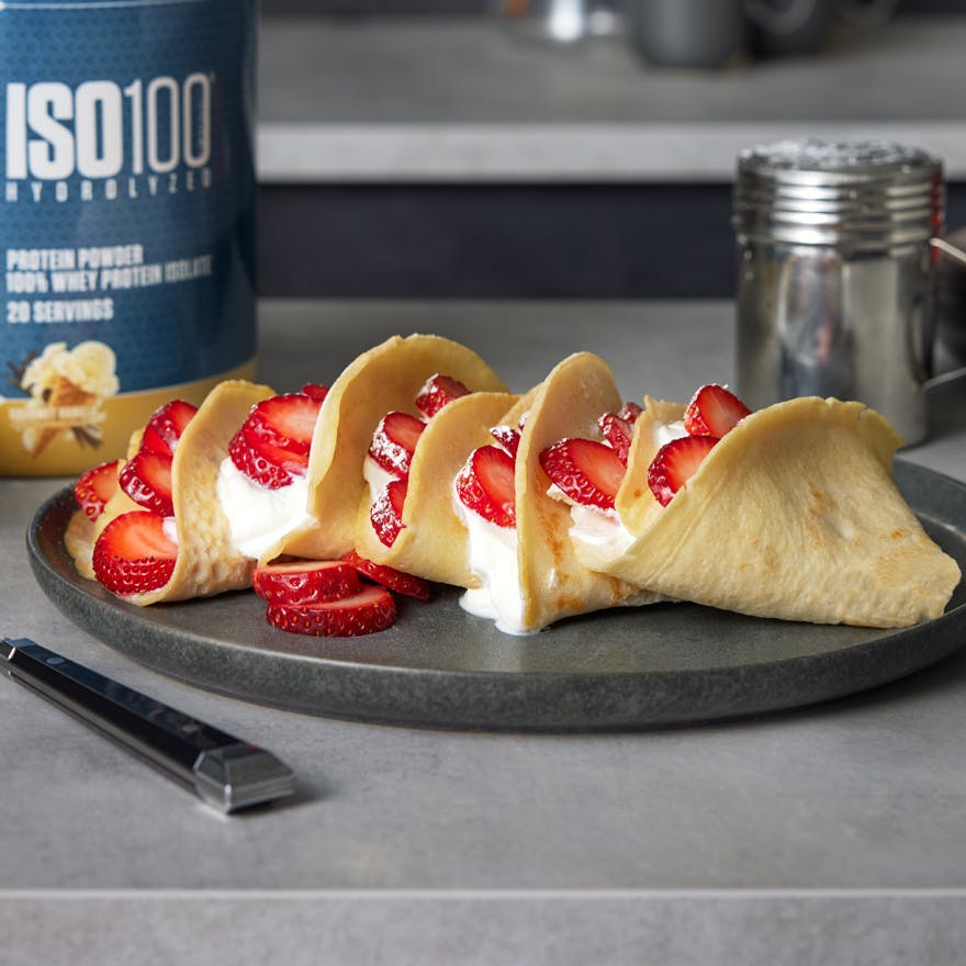 Five strawberry crepes on a plate with whipped cream. A tub of vanilla Dymatize ISO100 is on the table nearby