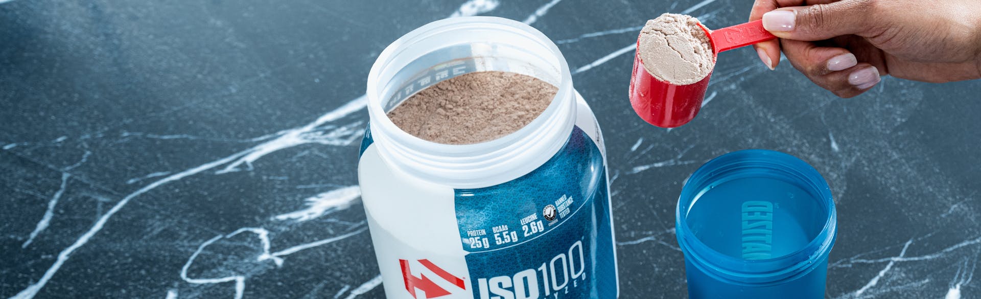 A single tub of Dymatize ISO100 open, with a hand pulling a scoop of powder out in a scooper.
