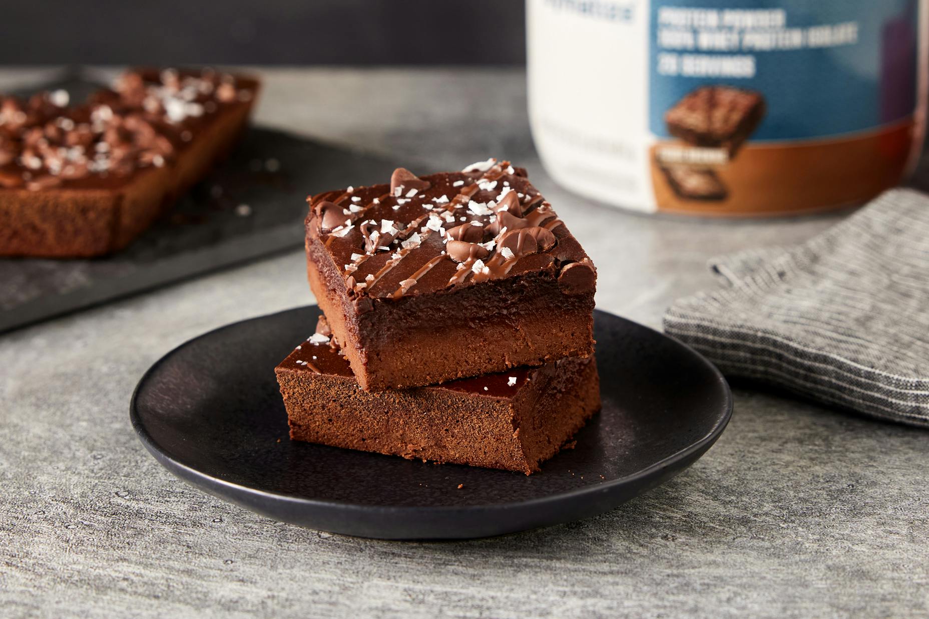 Protein Packed Brownies on a plate with a tub of Dymatize ISO100 Fudge Brownie Protein Powder